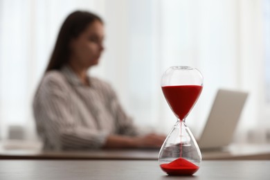 Photo of Hourglass with red flowing sand on table. Woman using laptop indoors, selective focus
