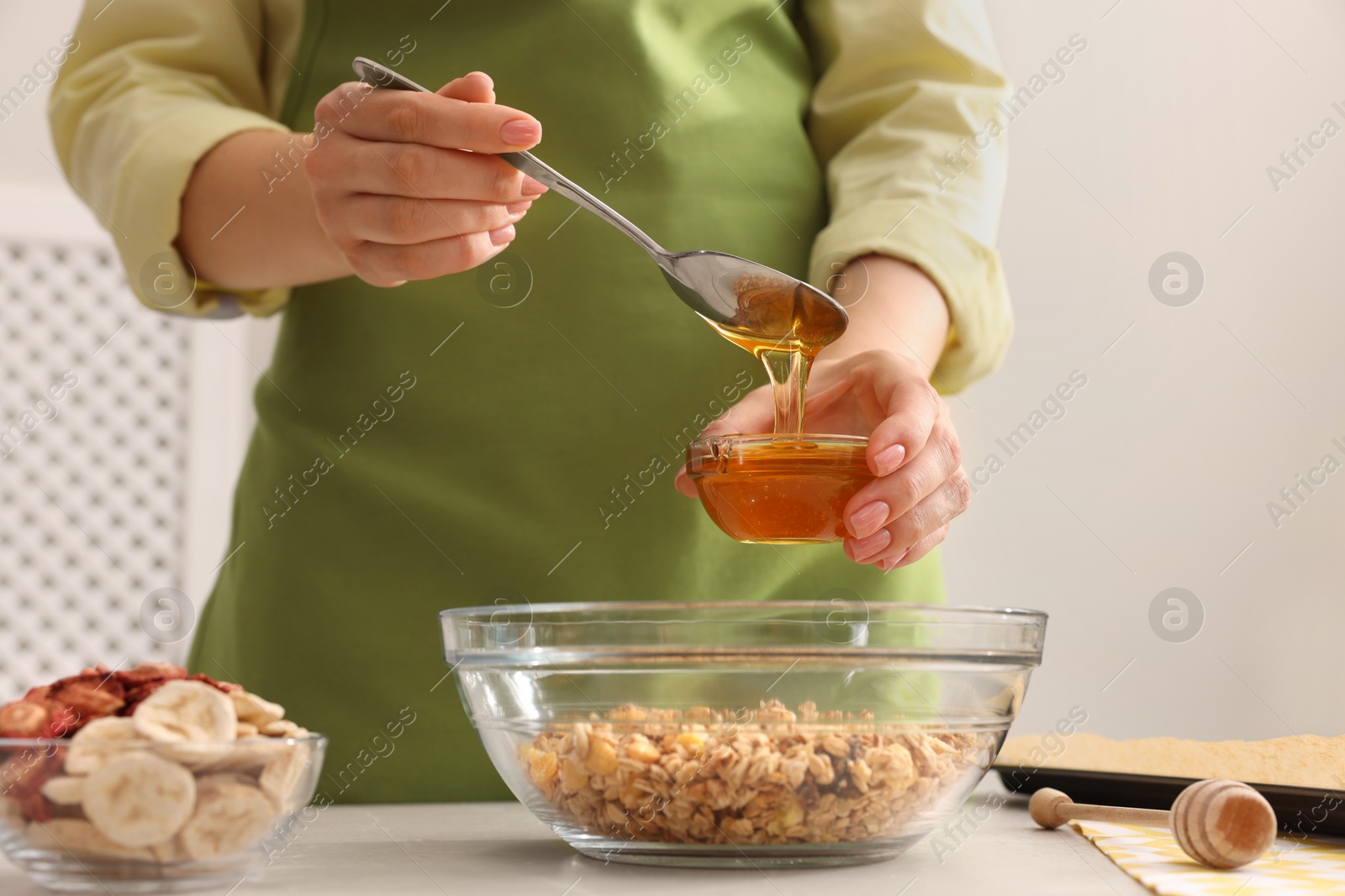 Photo of Making granola. Woman adding honey into bowl with mixture of oat flakes and other ingredients at light table in kitchen, closeup