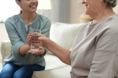 Photo of Young woman giving water to elderly lady indoors, closeup. Senior people care