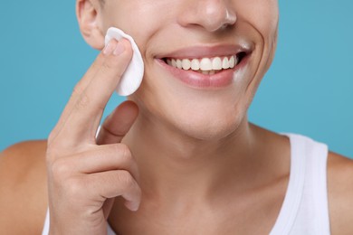 Man cleaning face with cotton pad on light blue background, closeup