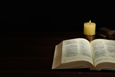 Church candle, Bible, cross and rosary beads on wooden table against black background, space for text