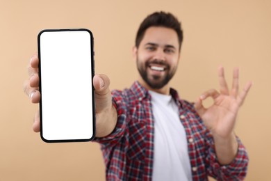 Photo of Young man showing smartphone in hand and OK gesture on beige background, selective focus. Mockup for design