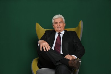 Happy mature businessman sitting in armchair against green background