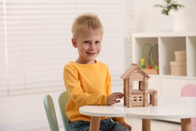 Cute little boy playing with wooden house at white table indoors. Child's toy