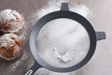 Photo of Sieve with sugar powder and muffins on grey textured table, flat lay