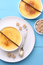Delicious creme brulee in bowls, vanilla pods, sugar cubes and spoon on light blue wooden table, flat lay