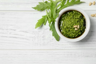 Bowl of tasty pesto and arugula on white wooden table, flat lay. Space for text