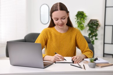E-learning. Young woman taking notes during online lesson at white table indoors