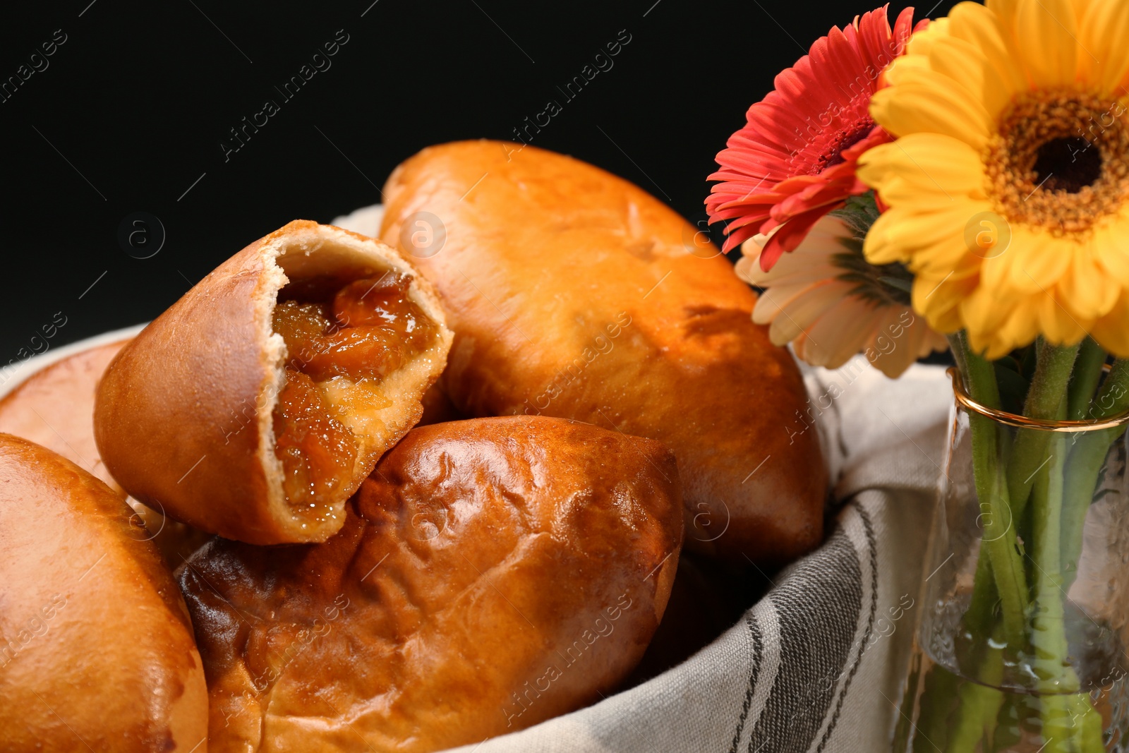 Photo of Many delicious baked patties and flowers on black background, closeup