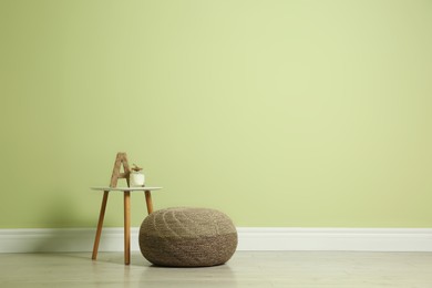 Photo of Comfortable knitted pouf and table with decor elements near light green wall indoors, space for text. Interior design