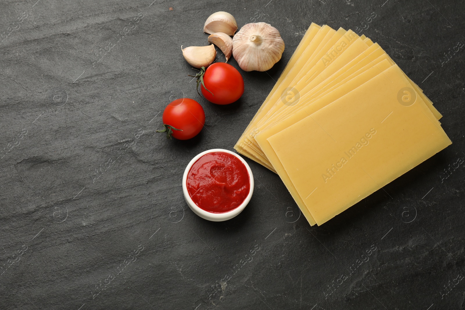 Photo of Cooking lasagna. Pasta sheets, tomato, garlic and ketchup on dark textured table, flat lay. Space for text