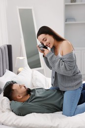 Happy young couple spending time in bedroom. Smiling woman taking photo of her boyfriend