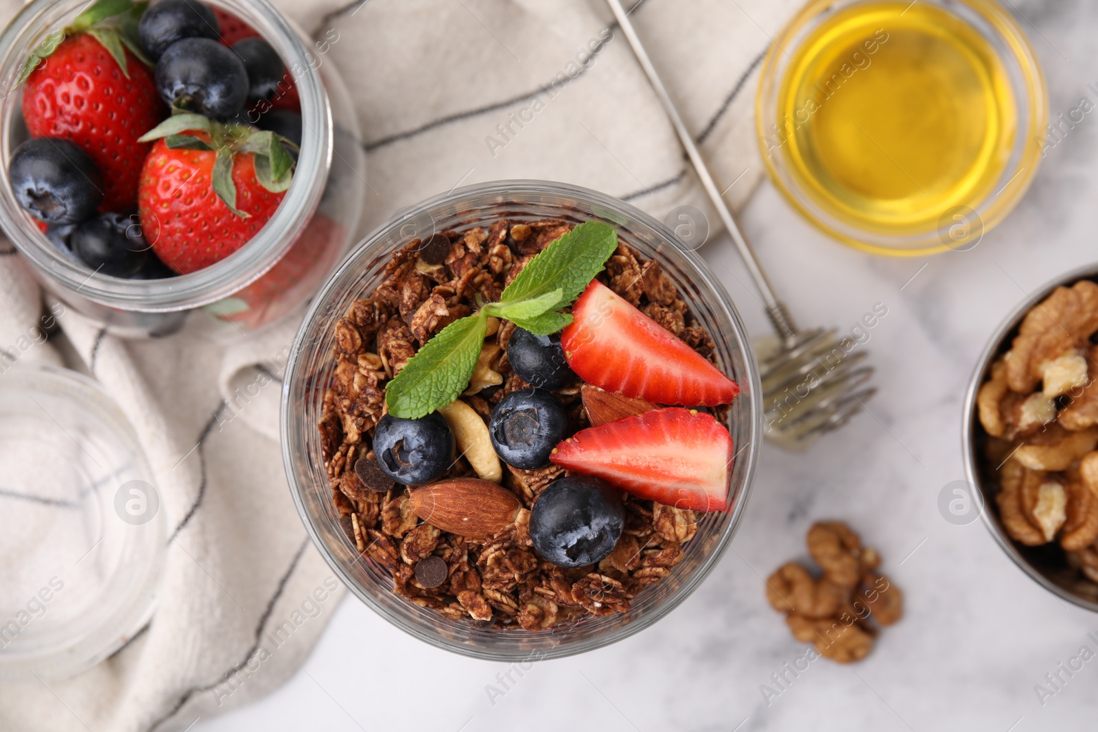 Photo of Tasty granola with berries, nuts, honey and whisk on white marble table, flat lay