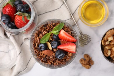 Photo of Tasty granola with berries, nuts, honey and whisk on white marble table, flat lay