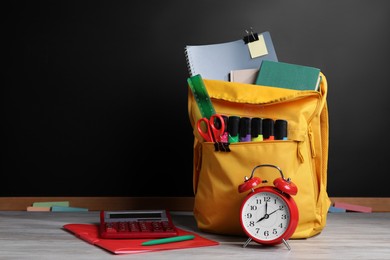 Photo of Backpack with different school stationery on white wooden table near blackboard