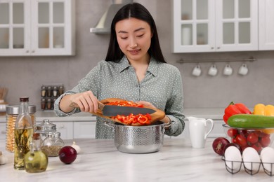 Photo of Cooking process. Beautiful woman adding cut bell pepper into pot in kitchen