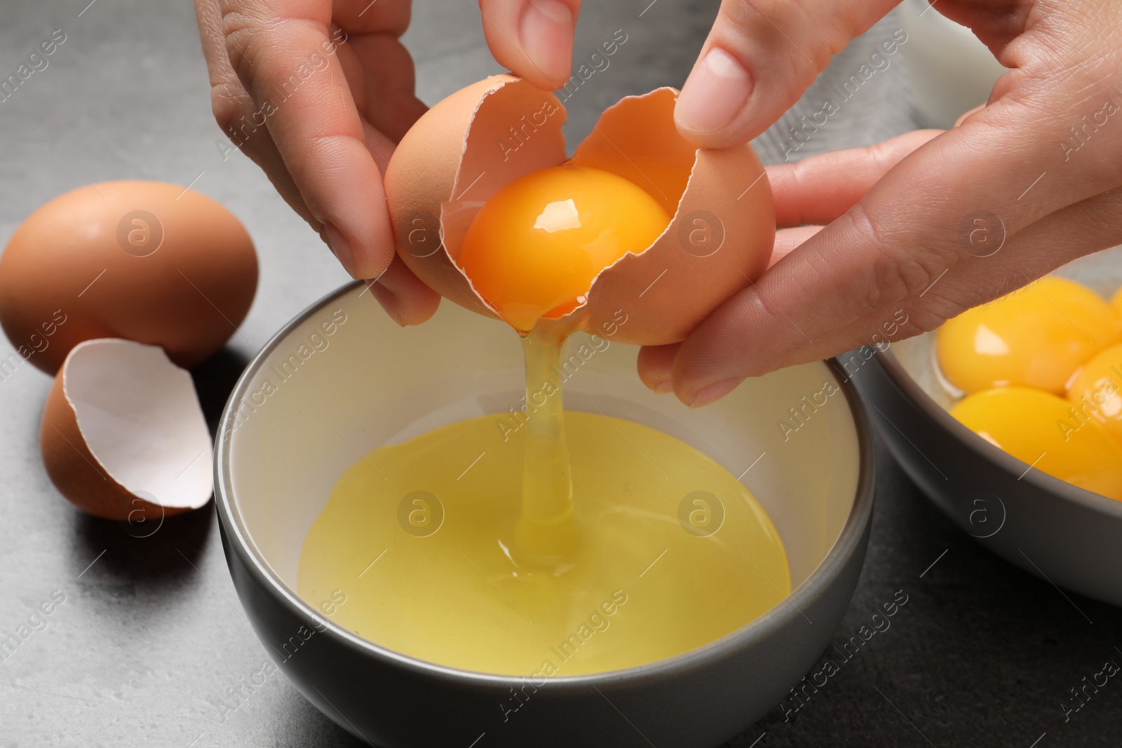 Photo of Woman separating egg yolk from white over bowl at grey table, closeup