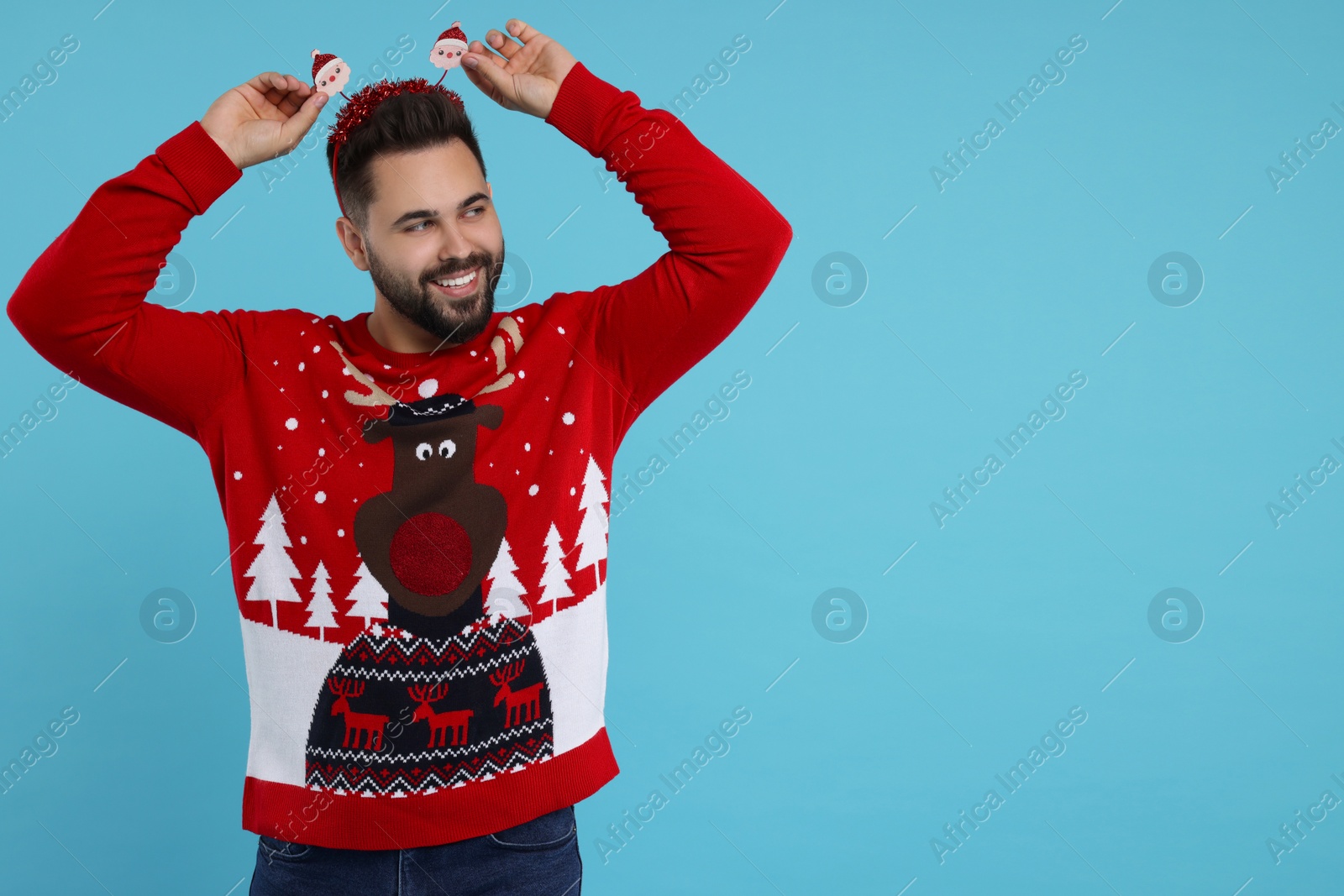 Photo of Happy young man in Christmas sweater and Santa headband on light blue background. Space for text