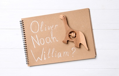 Photo of Notebook with different baby names and wooden dinosaur toy on white table, top view