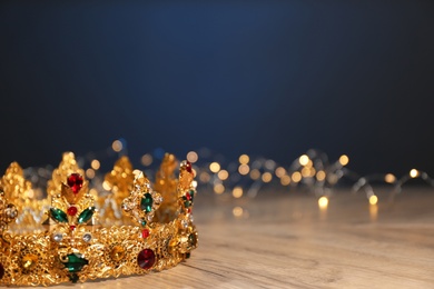 Photo of Beautiful golden crown with gems and fairy lights on wooden table, space for text. Fantasy item