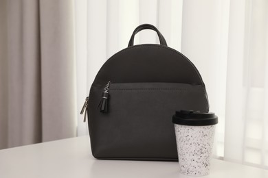 Stylish black backpack and cup of coffee on white table indoors