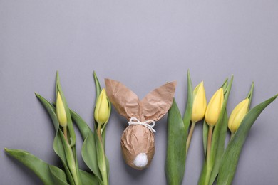 Photo of Easter bunny made of kraft paper and egg near beautiful tulips on grey background, flat lay. Space for text