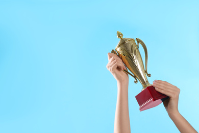 Photo of Woman holding gold trophy cup on light blue background, closeup. Space for text