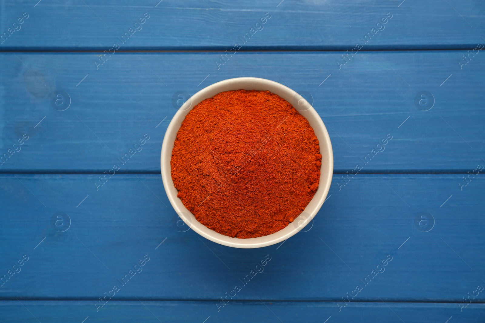 Photo of Bowl with aromatic paprika powder on blue wooden table, top view