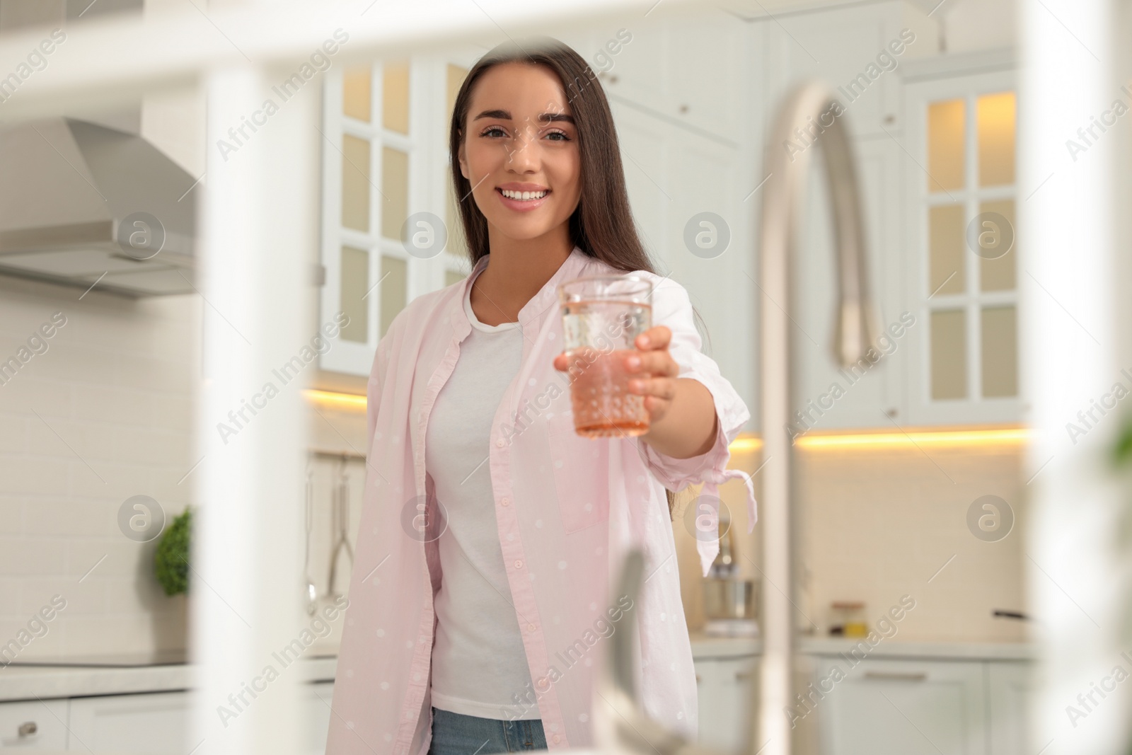 Photo of Young woman holding glass of pure water in kitchen, view through window