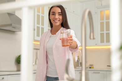 Young woman holding glass of pure water in kitchen, view through window