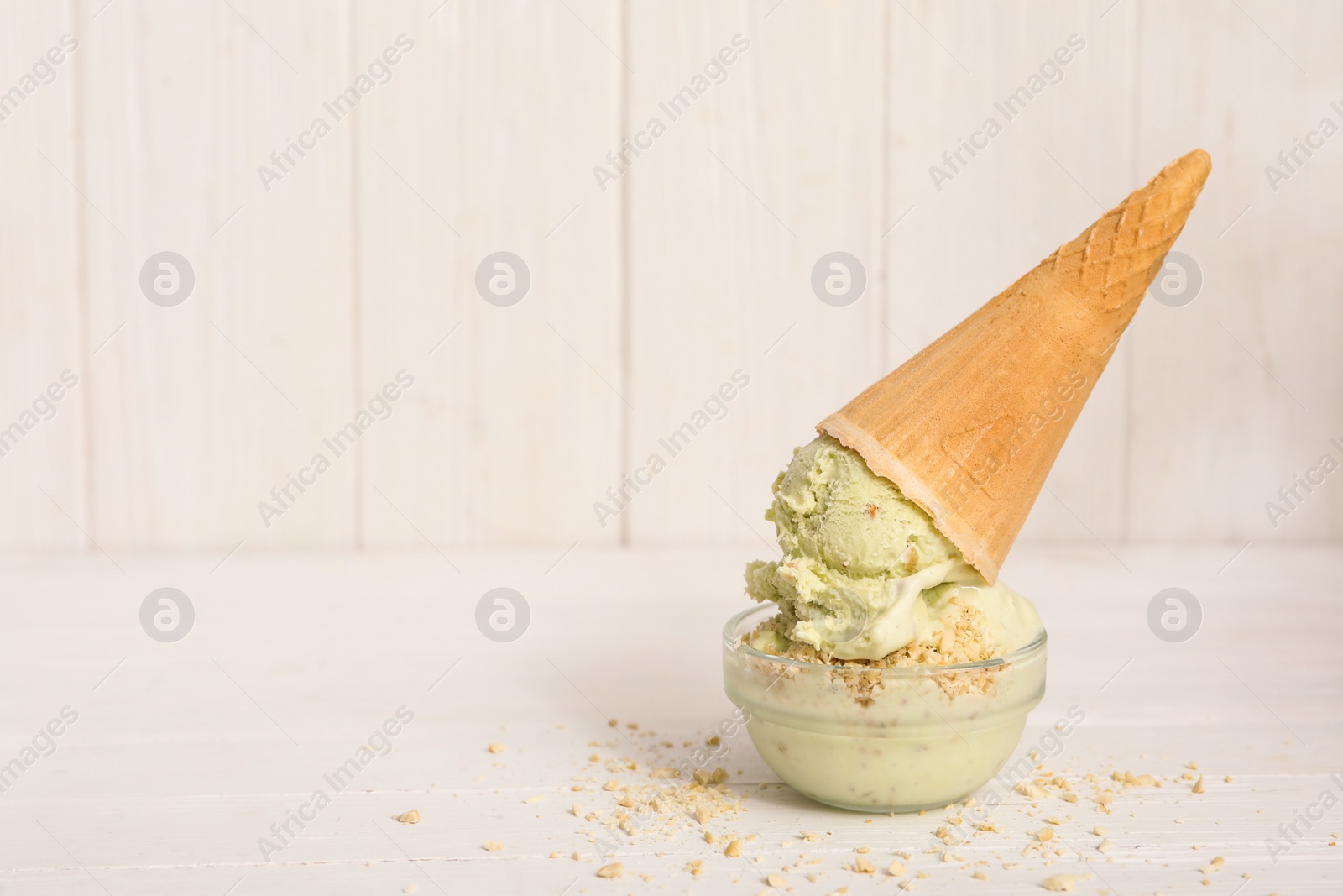 Photo of Delicious pistachio ice cream in wafer cone with chopped nuts on white wooden table. Space for text