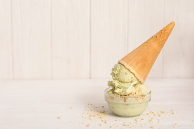 Photo of Delicious pistachio ice cream in wafer cone with chopped nuts on white wooden table. Space for text