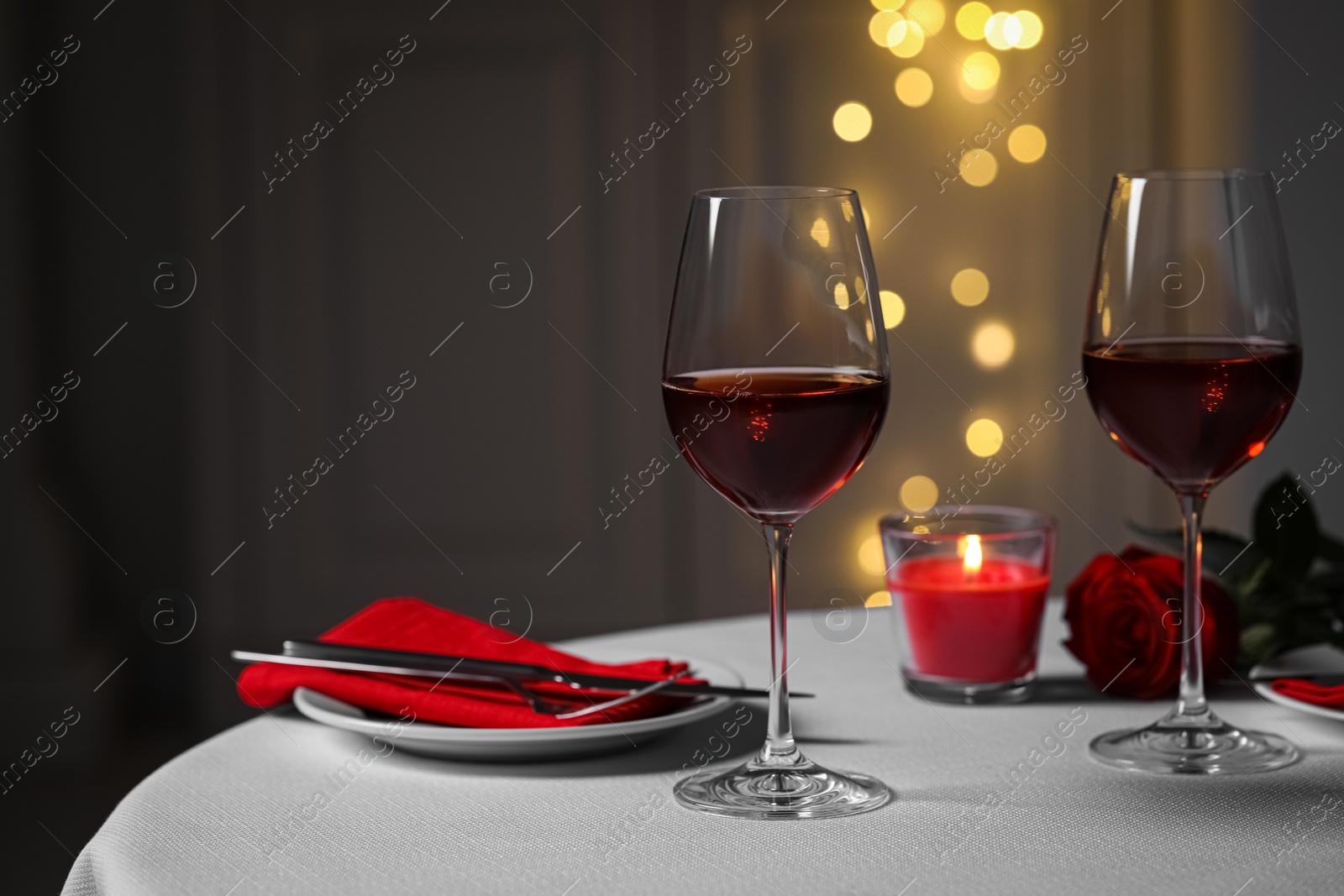 Photo of Romantic table setting with glasses of red wine, rose flower and burning candle against blurred lights, space for text