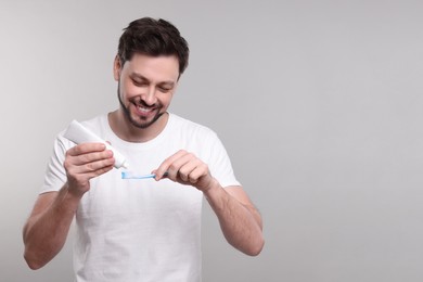 Photo of Happy man squeezing toothpaste from tube onto toothbrush on light grey background. Space for text