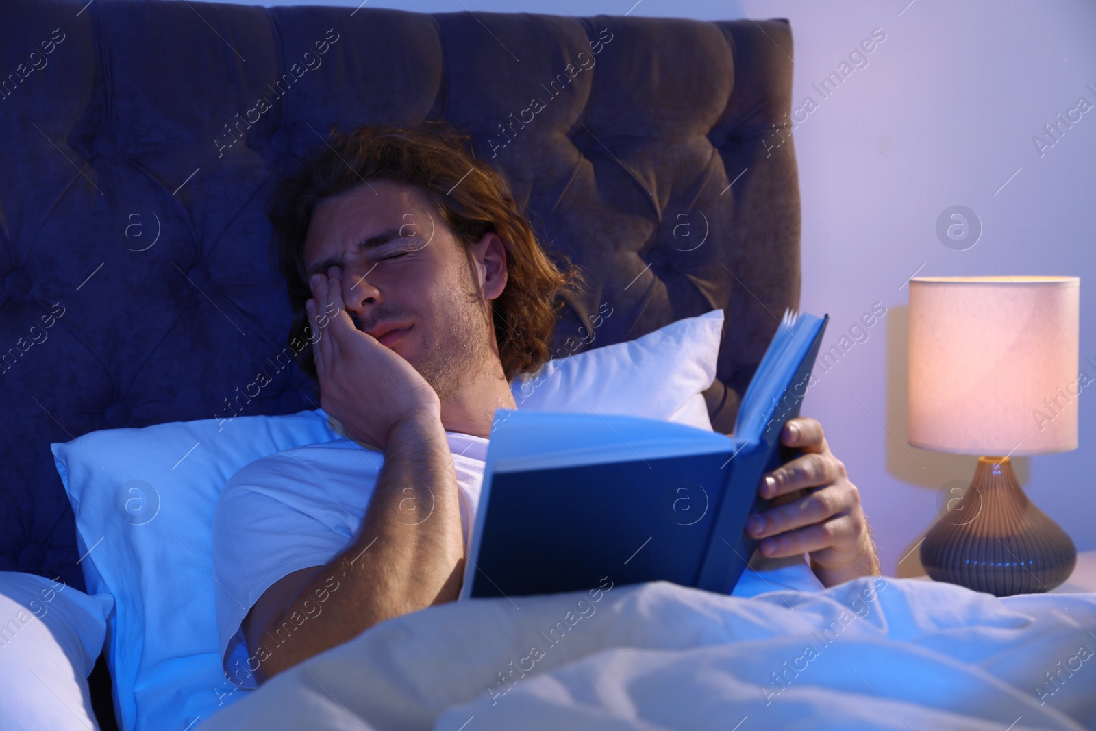 Photo of Sleepy young man with book in dark room at night. Bedtime