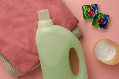 Photo of Bottle of fabric softener, stacked clean clothes, laundry capsules and detergent powder on pink background, flat lay