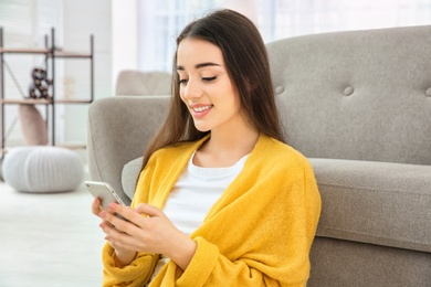 Photo of Attractive young woman using mobile phone sitting near sofa at home