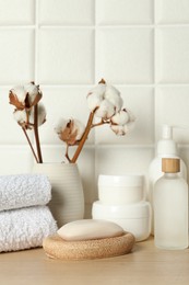 Photo of Different bath accessories, personal care products and cotton flowers in vase on wooden table near white tiled wall