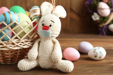 Photo of Toy bunny in protective mask near dyed eggs on wooden table. Easter holiday during COVID-19 quarantine