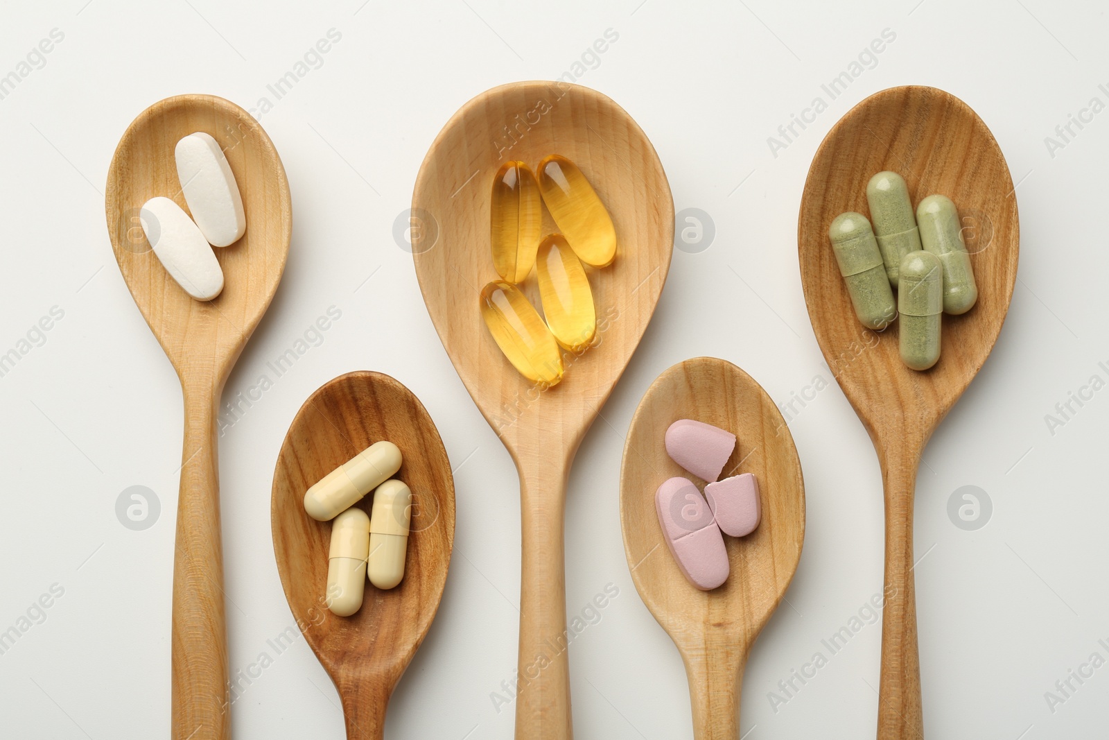 Photo of Vitamin capsules in wooden spoons on white background, flat lay