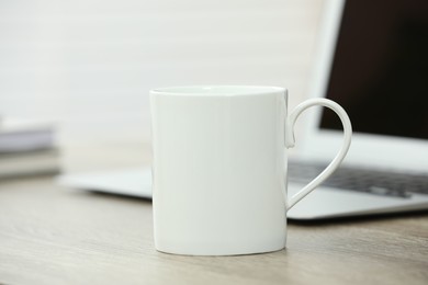 Photo of White ceramic mug and laptop on wooden table indoors