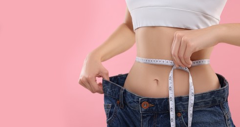 Photo of Slim woman wearing big jeans and measuring waist with tape on pink background, closeup. Weight loss
