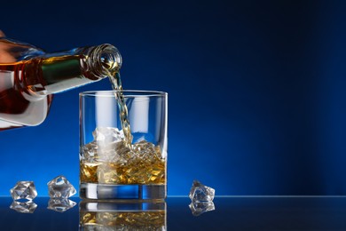 Pouring whiskey into glass with ice cubes at table against dark blue background, space for text