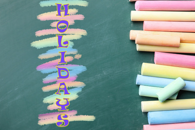 Image of Word HOLIDAYS and pieces of color chalk on greenboard, flat lay