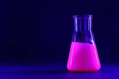 Photo of Laboratory flask with luminous liquid on dark blue background, space for text