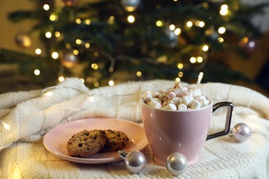 Photo of Cup of tasty cocoa with marshmallows and cookies on knitted plaid near Christmas tree indoors