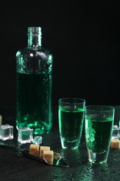 Photo of Absinthe in shot glasses, spoon, brown sugar and ice cubes on gray table against dark background. Alcoholic drink