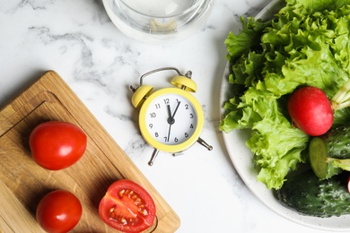 Photo of Alarm clock and vegetables on white marble table, flat lay. Meal timing concept