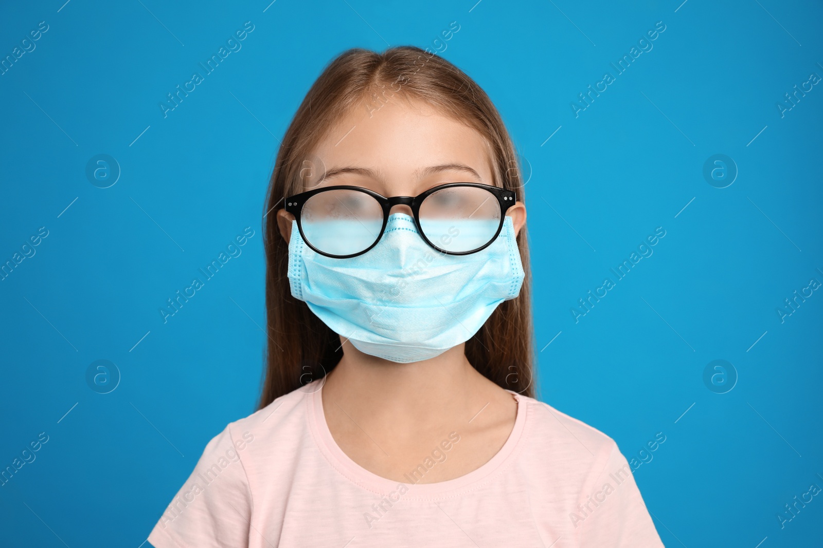 Photo of Little girl with foggy glasses caused by wearing disposable mask on blue background. Protective measure during coronavirus pandemic