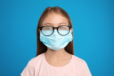 Little girl with foggy glasses caused by wearing disposable mask on blue background. Protective measure during coronavirus pandemic